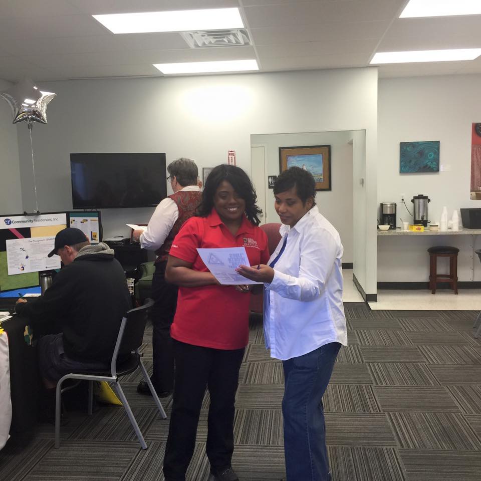 Two black women standing in the community recovery center looking at a piece of paper. Other are filling out papers in the background. Arundel House of Hope connects people to resources and skill developement. 