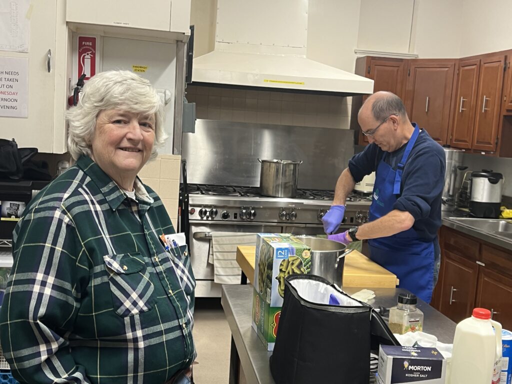 A volunteer and board member smiling while standing in a kitchen. In the background a volunteer is preparing an evening meal for people experiencing homelessness. 