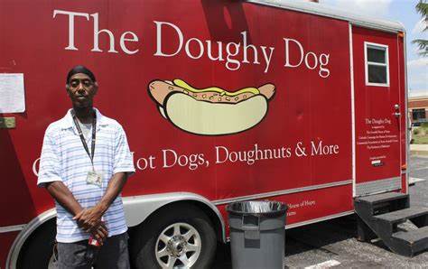 A young black man standing in front on a food truck that says The Doughy Dog. Hot Dogs, Doughnuts, and More. The food truck has a large picture of a hot dog. The young man has a cap and is clasping his hands in front of him. 