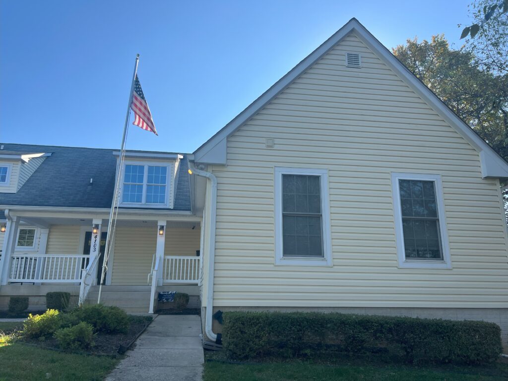 The Patriot house is yellow and large. There is a flag pole displaying the flag in front of the house. 