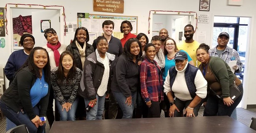  A large group of young people are standing in front of a table with big smiles. The group has black, white, and Asian people in it. Volunteers help us meet our mission to end homelessness 