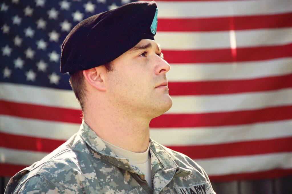 A man in a beret and camo uniform that says Army is looking off to the distance. There is an American flag in the background. Veterans can have homelessness and a variety of complex needs.