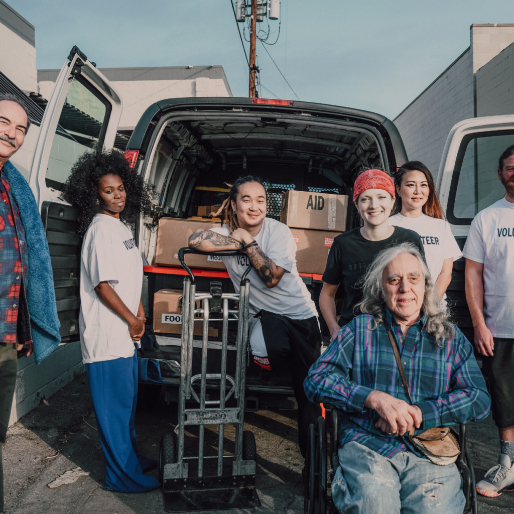 Volunteers standing around a moving van. An older man with white hair is sitting in a wheel chair. Volunteers help people who were homeless move into housing. 