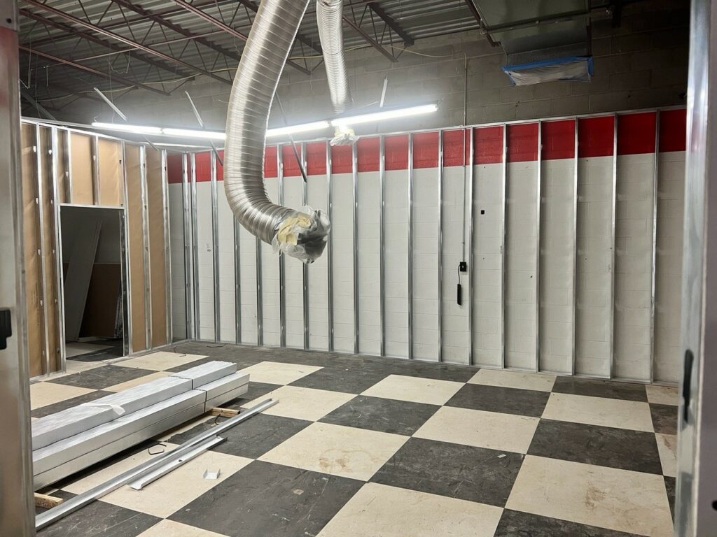 A room under construction. The floor tile is white and black checkers. The wall has no dry wall and you can steel beams. There is a duct hanging down. 