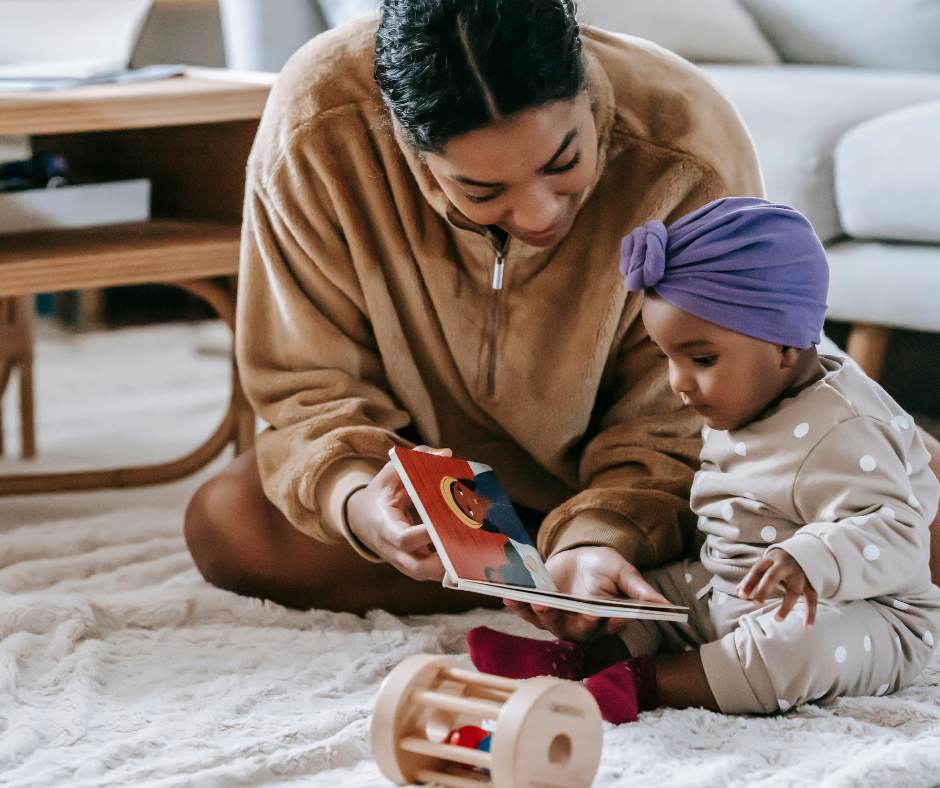 A black mom and small toddler sitting on the floor. There is a toy on the floor and the mom is hunched over holding a board book for the baby. Mom and baby are reading together. The Family Project supports both mom and children. 