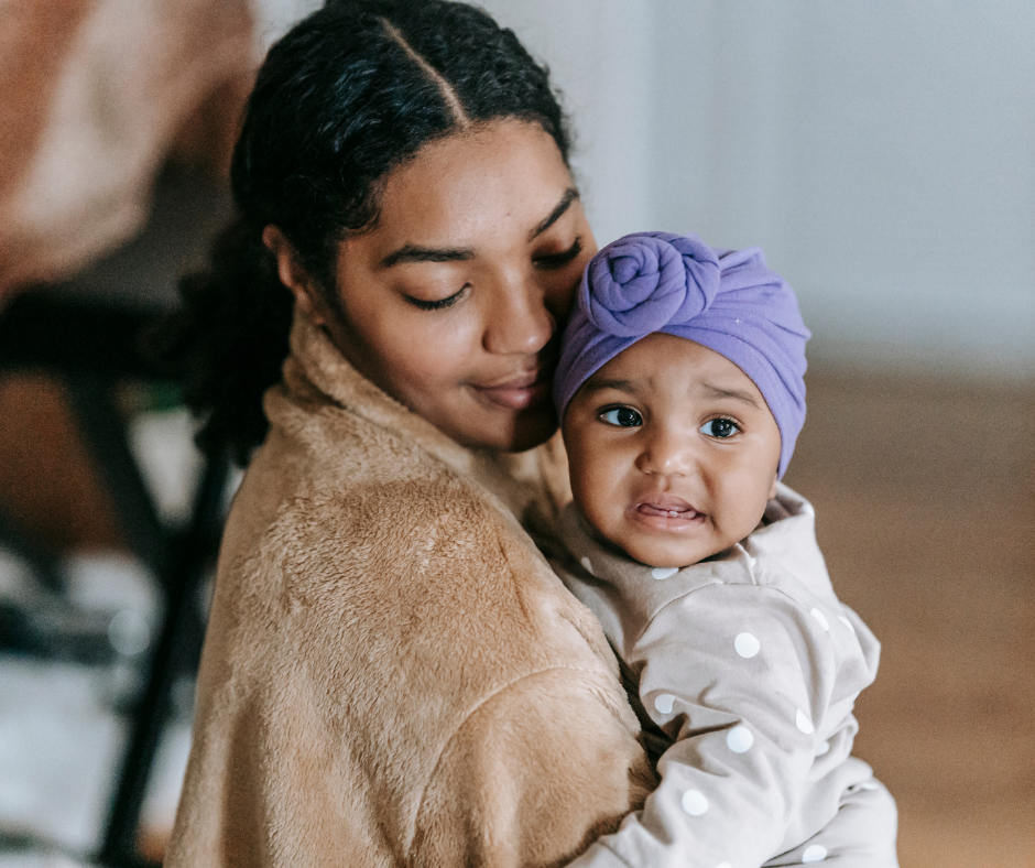 a young black mother holding her daughter with a smile on her face. The baby has a purple scarf on her head and looks like she might start crying. Homeless mothers in recovery benefit from support and a stable home.