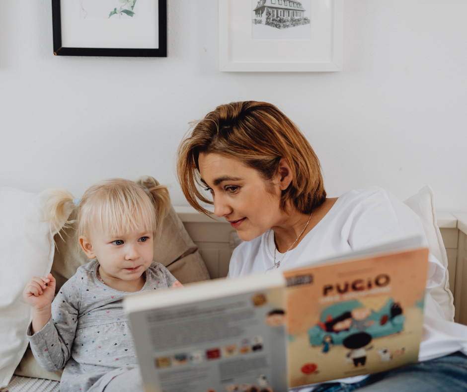 a white women with short hair reading a book to a small child. The child is white and has blond pig tails. Children are healthier if they have stable housing. 
