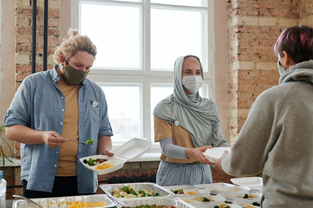Two women serve warm lunches. One woman is blond and has on a blue shirt over a white t-shirt. The other women wear a blue hijab. Volunteers often share meals and social with people experiencing homelessness in our day center. 