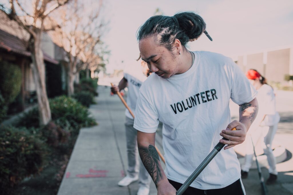 An Asian man in a white t-shirt and long black hair tied into a bun is spreading mulch. Volunteers help keep our community housing in great shape. 