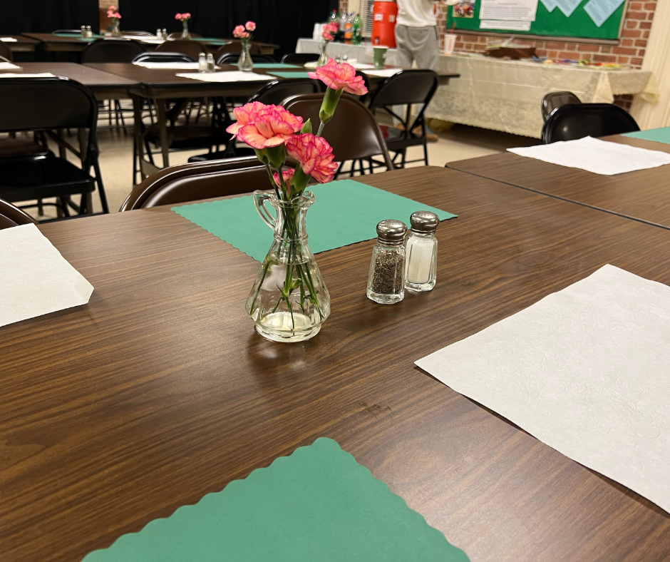 A table set with green and white paper placemats. There is a vase of fresh flowers and salt and pepper on the table. Winter Relief volunteers and guests eat dinner together. 