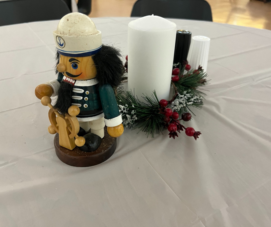 A table with a white table cloth. The center piece is a white handle with greenery at the base with a small nutcracker statue of a sailor at a steering wheel. Each winter relief host cite decorates and adds comforting touches to their buildings when hosting people experiencing homelessness. 
