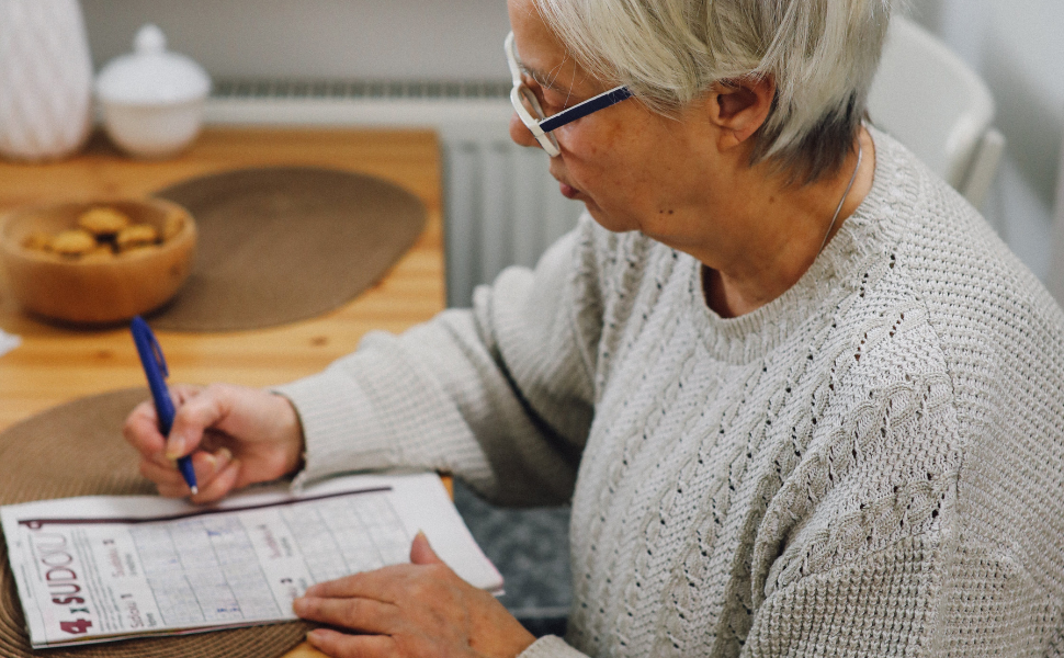 An older white woman with short hair sits at a kitchen table working on a crossword puzzle. people who are stabile housed can peruse personal interests and recreation.  
