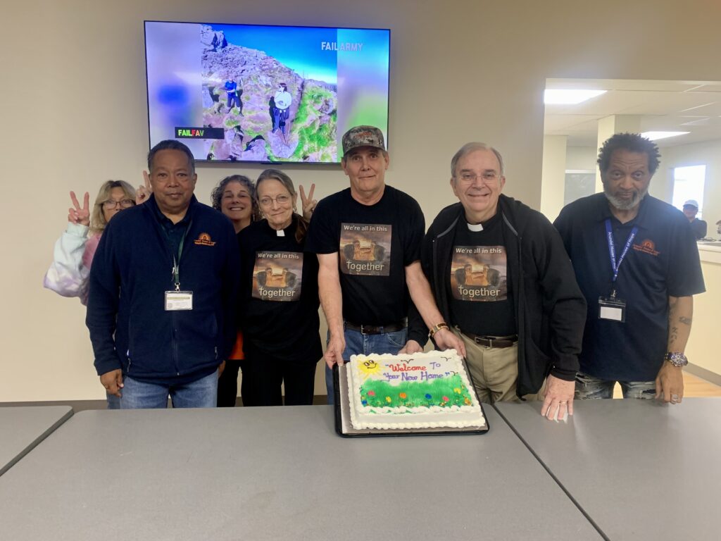 A group of people standing around a table holding a large cake that says welcome to your new home. The people are smiling 