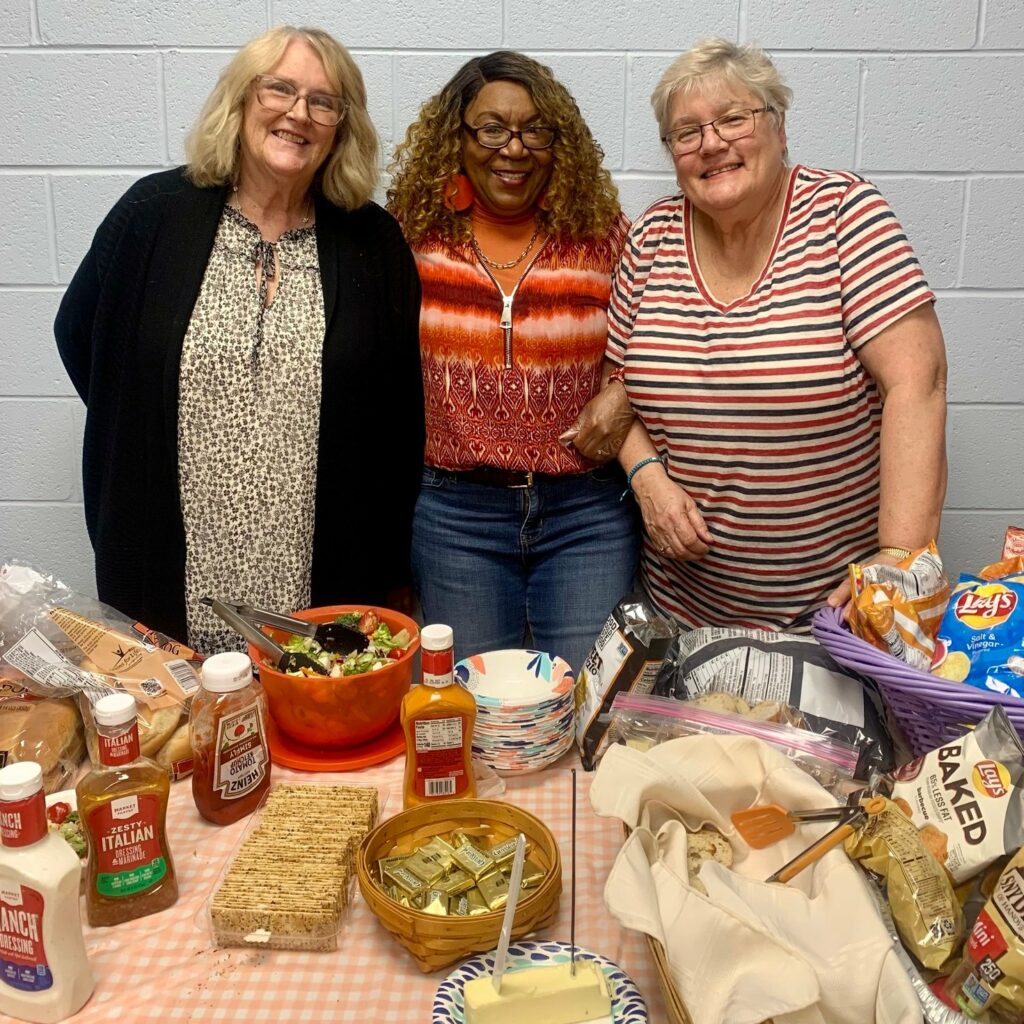 Solley volunteers are always ready to serve. Three ladies are standing behind a table full of food and have their arms hooked together. They are happy. 